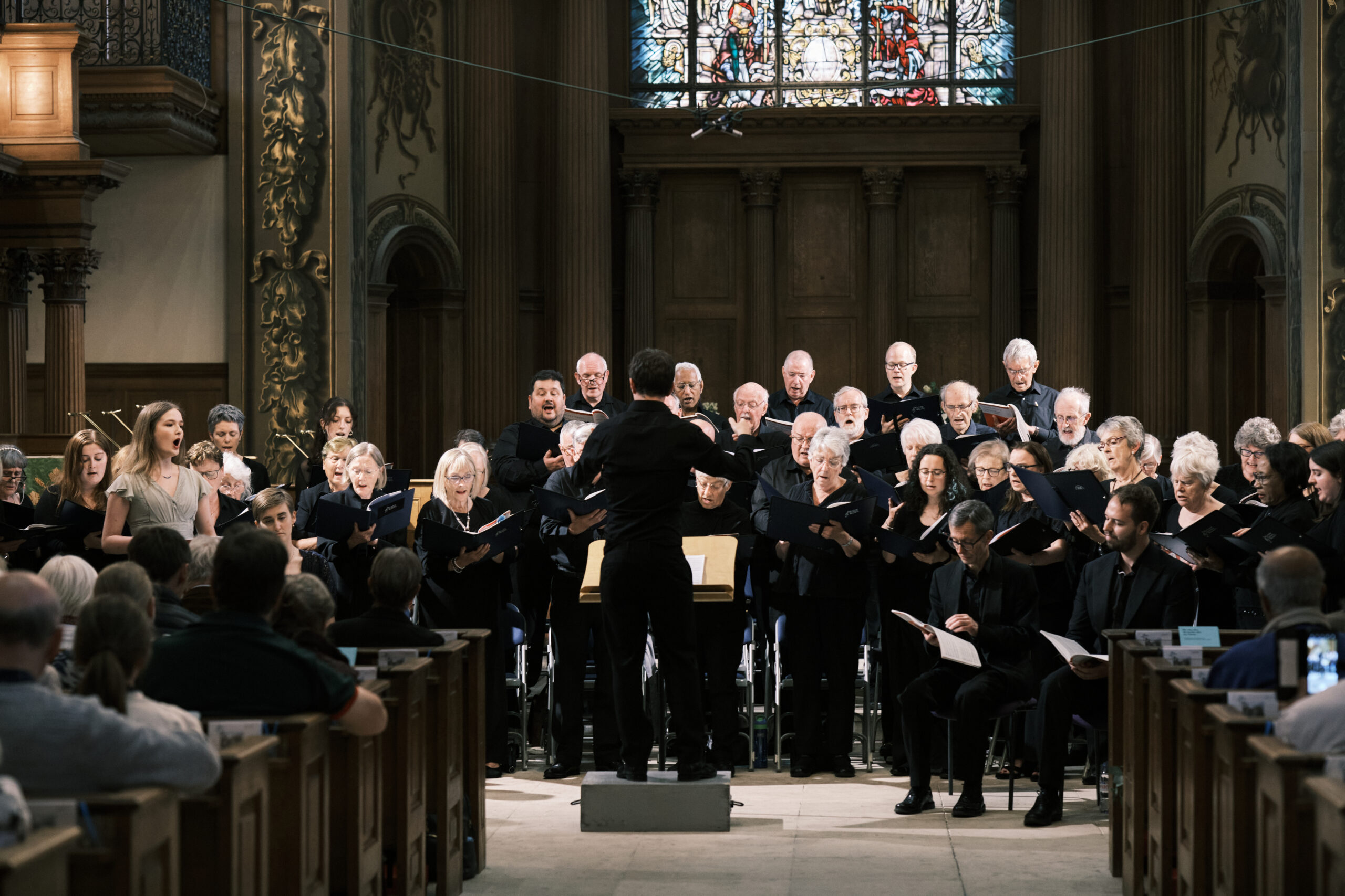 A wide image taken from the back of the church during the Autumn 2024 ECS concert. Max Barley is standing in the middle of the image on a block with his back to the camera conducting the choir. The choir are holding music folders and singing.