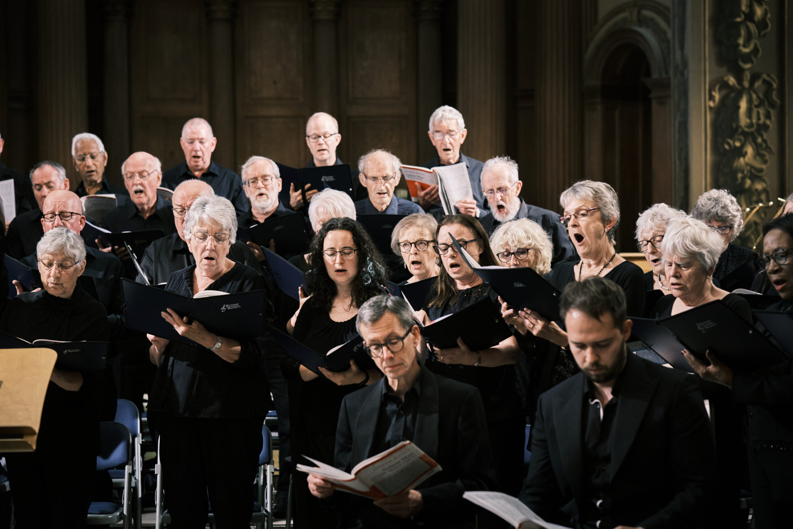 An image of 23 ECS members holding music folders and singing during the ECS Autumn 2024 concert. The photo is made up of tenors, basses and altos. At the front of the image two professional soloists are in view following along the music.