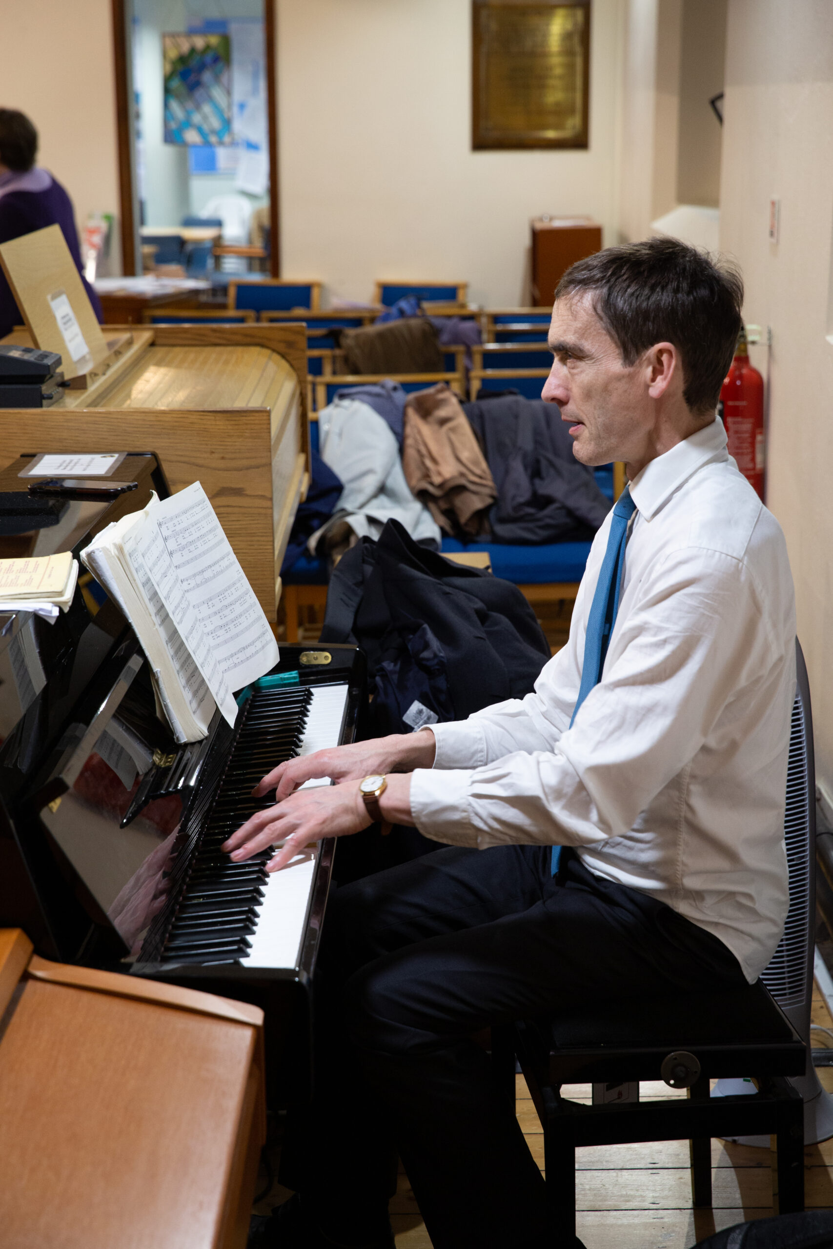 Photo of Andrew Lenon, Accompanist of Eltham Choral Society, playing the piano at a rehearsal. Andrew is wearing a shirt and tie.