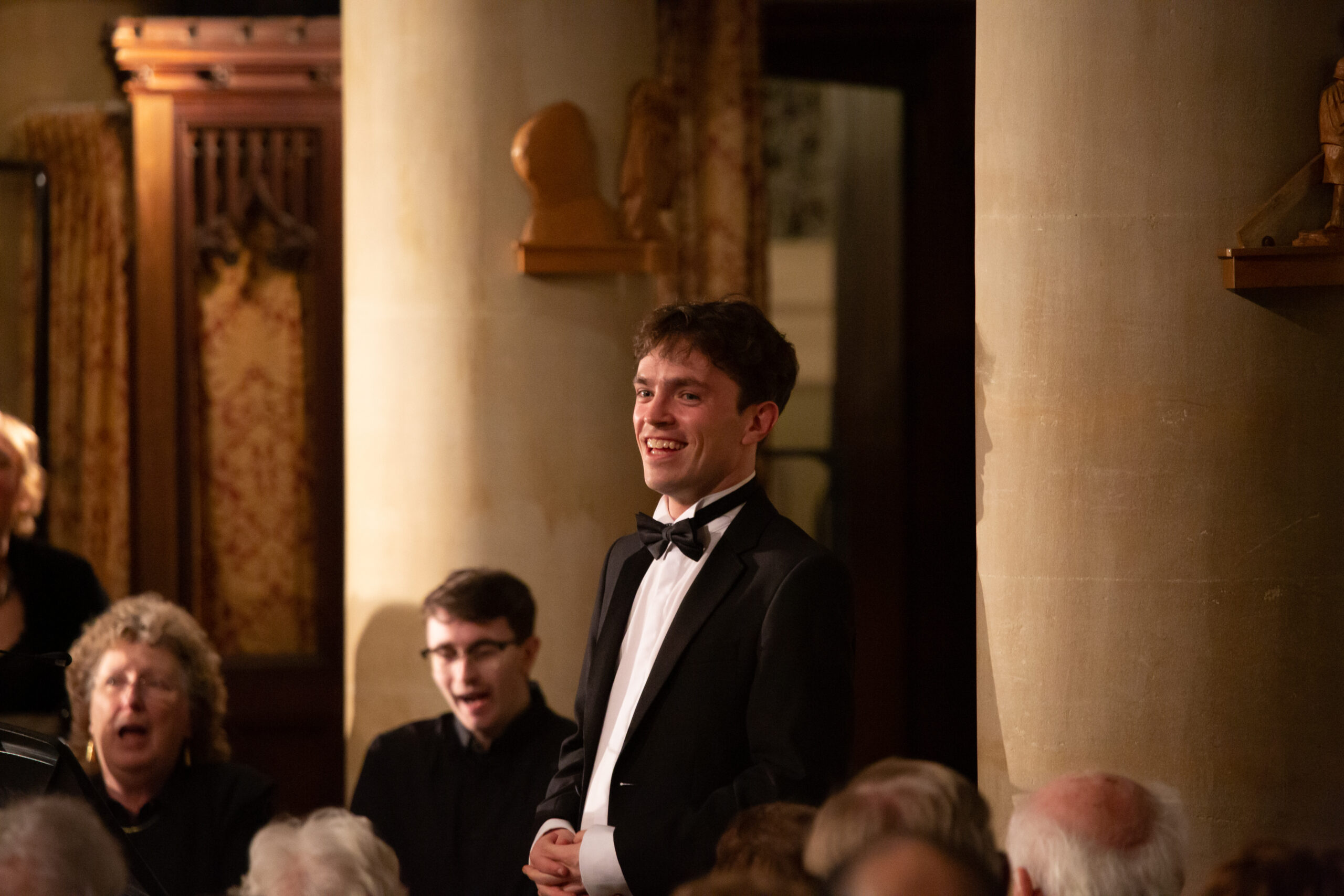 Photo of Max Barley, Conductor of Eltham Choral Society, wearing black tie at an ECS concert. Max is smiling at the audience with a few members of the choir visible behind him.
